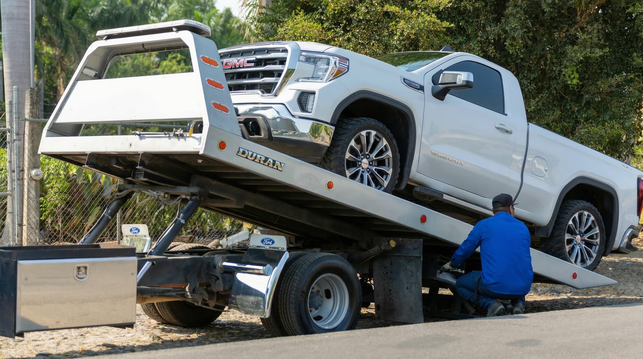Prime O Towing flatbed loading a white GMC Sierra Denali while a Prime O Towing driver secures the tie-downs